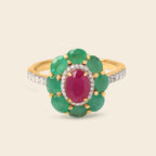 A yellow gold ring featuring a flower-shaped cluster with green emerald-cut stones, a central oval pinkish-red ruby-cut stone, and a halo of small clear stones, set against a light beige background.