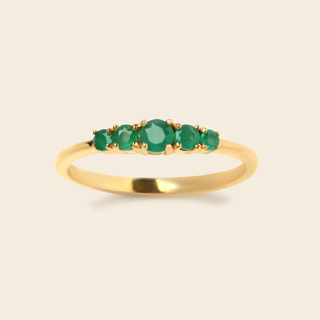 A yellow gold ring featuring five round-cut green emerald-cut gemstones in a row, set against a solid light beige background.