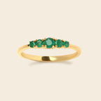 A yellow gold ring featuring five round-cut green emerald-cut gemstones in a row, set against a solid light beige background.