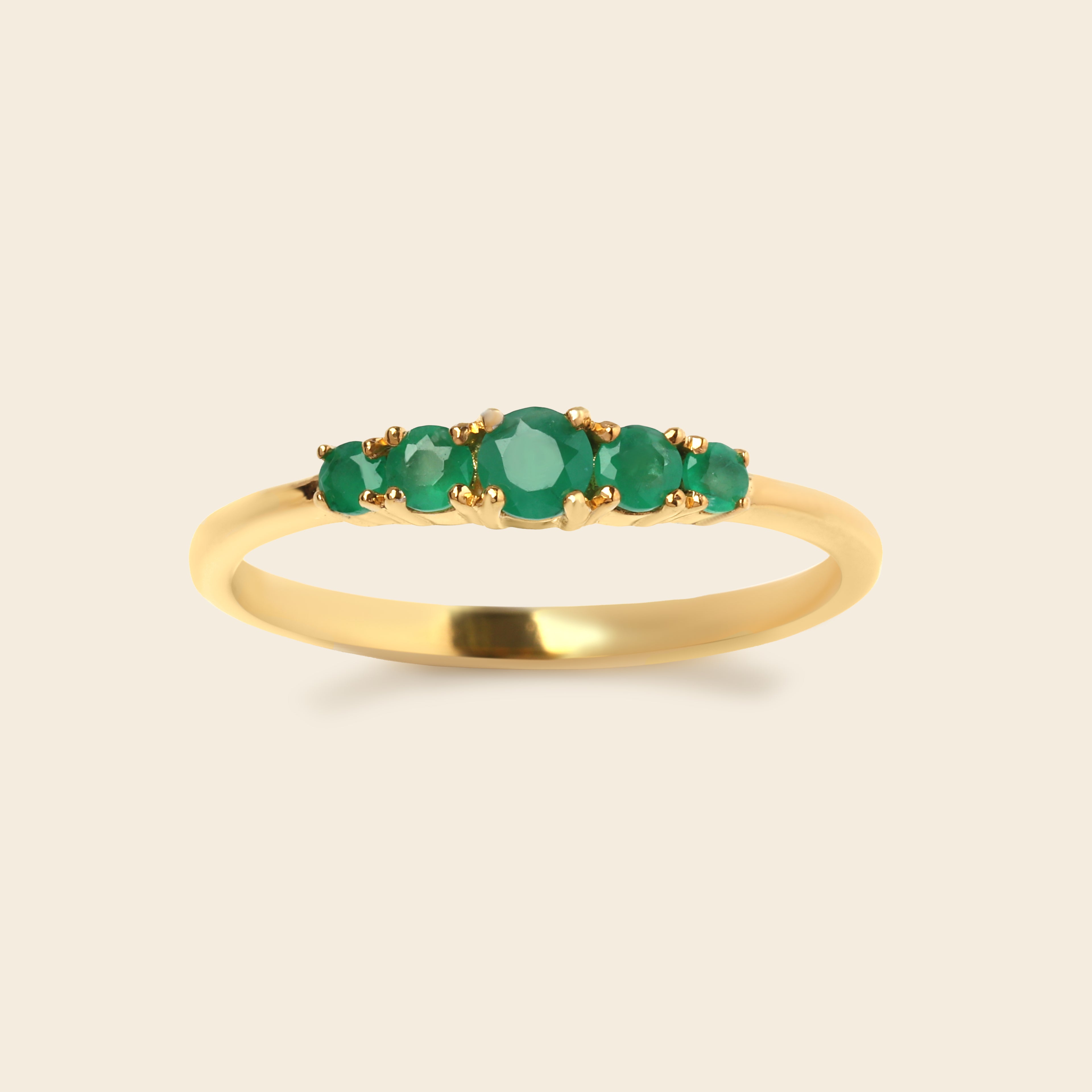 A yellow gold ring featuring five round-cut green emerald-cut gemstones in a row, set against a solid light beige background.