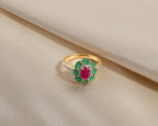 A yellow gold ring featuring a central oval ruby gemstone surrounded by a halo of green emerald-cut stones, with a pavé-set silver band, displayed on a draped cream satin fabric.