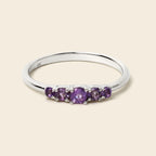 A silver five-stone ring featuring round purple amethyst gemstones, set against a solid light beige background.