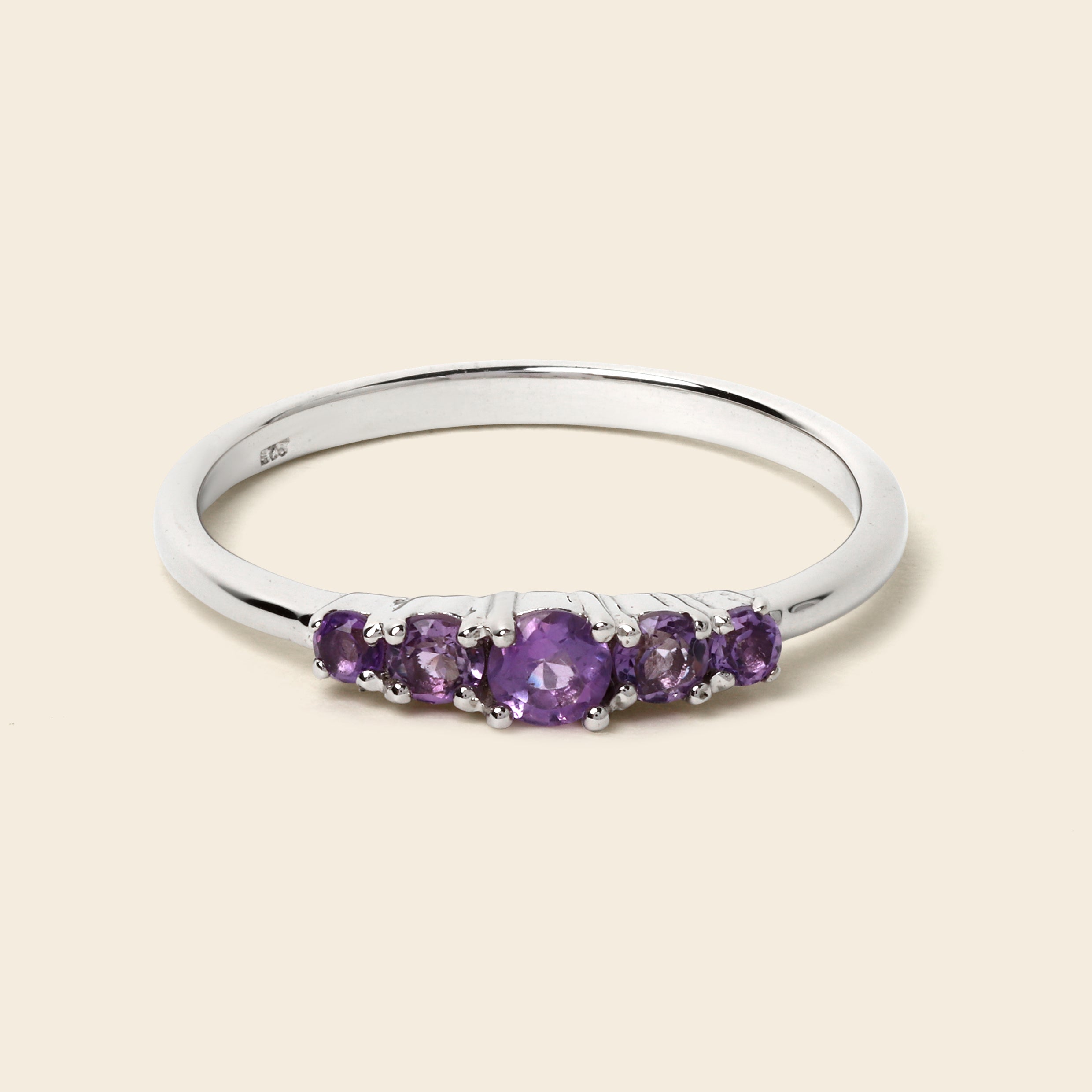 A silver five-stone ring featuring round purple amethyst gemstones, set against a solid light beige background.