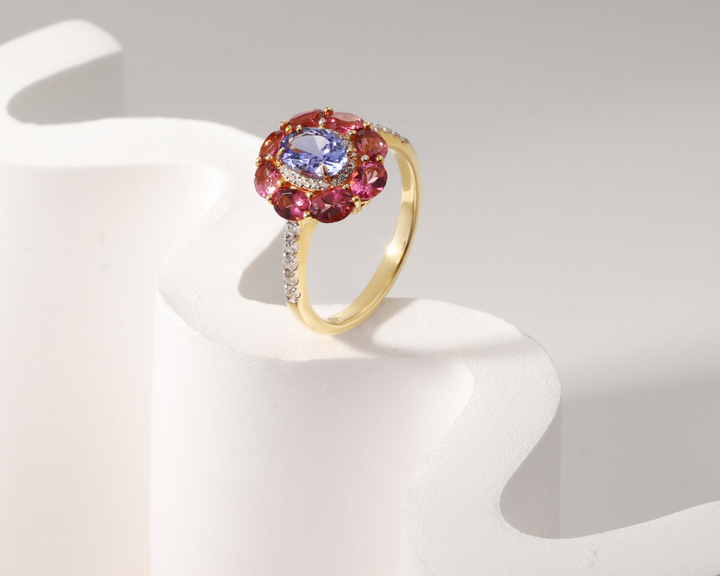 A yellow gold ring with a central oval blue gemstone surrounded by a halo of oval pink gemstones, accented with small clear stones on the band, displayed on a white sculptural stand.