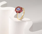 A yellow gold ring with a central oval blue gemstone surrounded by a halo of oval pink gemstones, accented with small clear stones on the band, displayed on a white sculptural stand.
