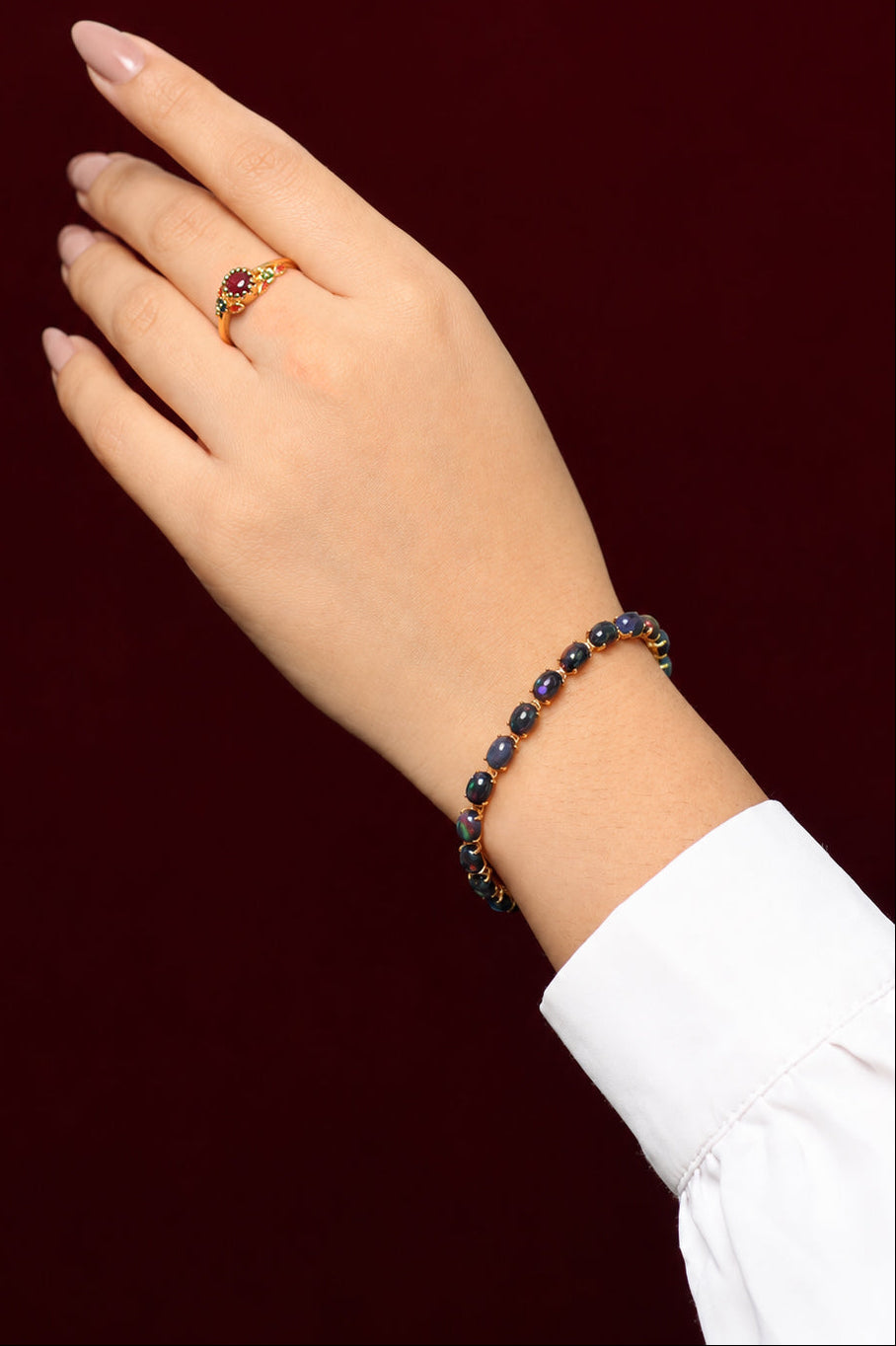 A close-up of a person's hand and wrist wearing a gold ring with a dark red oval stone and a matching beaded bracelet with dark blue and gold beads, set against a solid dark burgundy background.