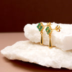 A pair of gold dangle earrings with green leaf-shaped stones, displayed on stacked white rock slabs against a dark brown background.