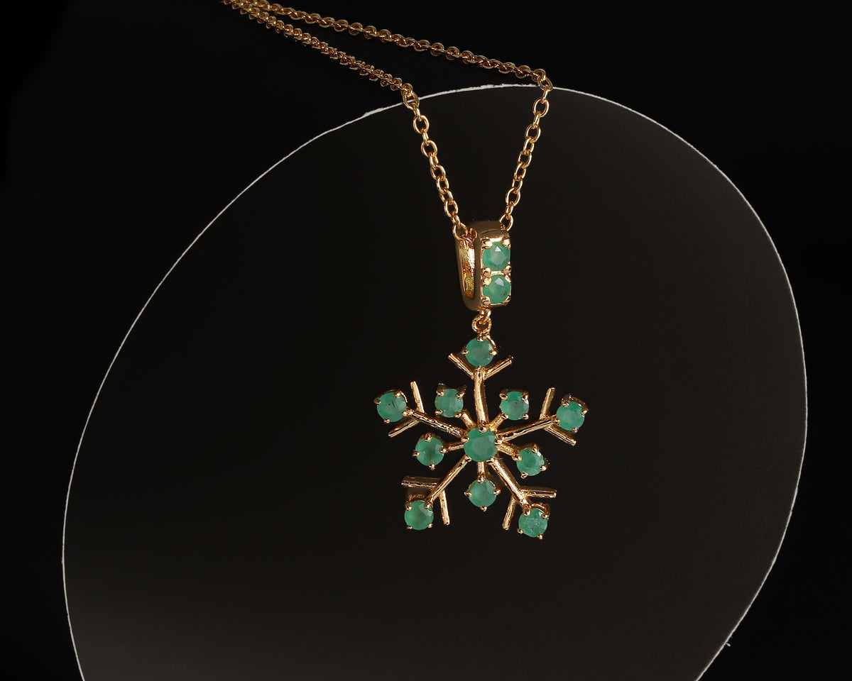 A gold snowflake pendant necklace with green gemstone accents, displayed on a clear circular stand against a black background.