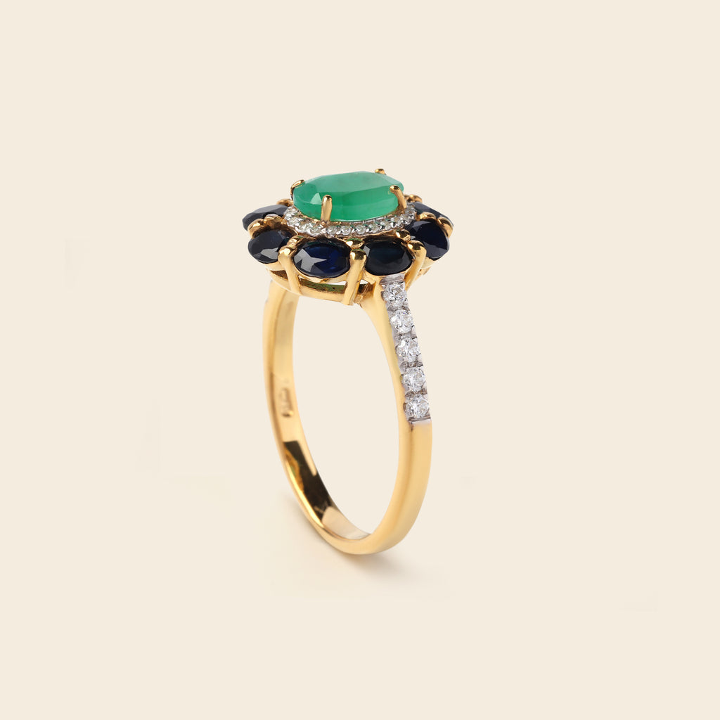 A yellow gold ring featuring a central oval green emerald-cut gemstone, surrounded by a halo of round dark blue gemstones and a pavé band of clear stones, set against a solid cream background.