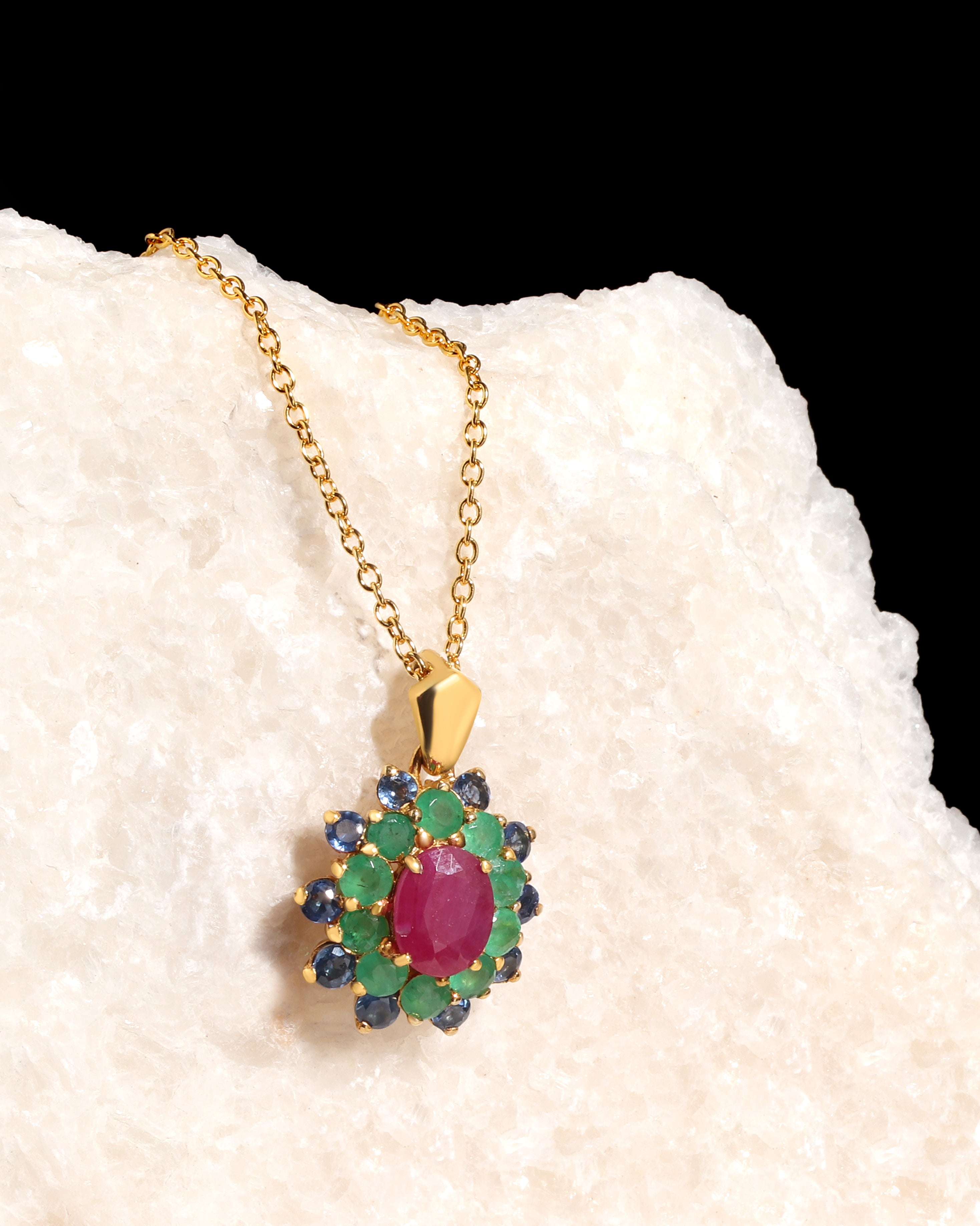 A gold pendant necklace featuring a central oval pinkish-red gemstone surrounded by green and dark blue round gemstones, displayed on a textured white stone against a black background.