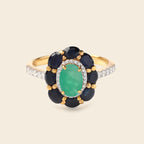A yellow gold ring featuring a central oval green emerald-cut stone surrounded by a halo of round dark blue sapphires and small clear accent stones on a light beige background.
