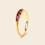 A gold ring featuring a row of round-cut pinkish-red gemstones, shown on a solid light beige background.