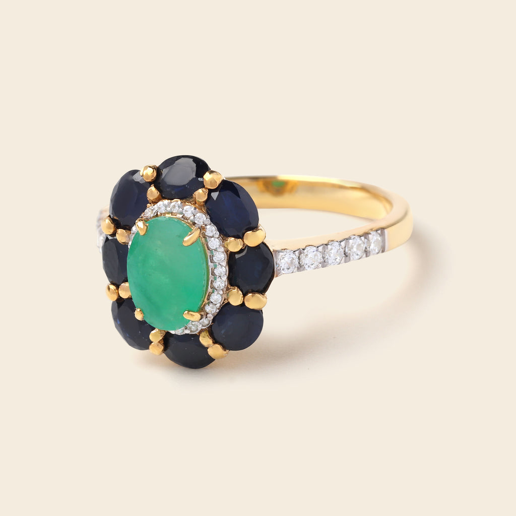 A yellow gold ring featuring a central oval green emerald-cut stone surrounded by a halo of dark navy blue stones, accented with small clear pavé diamonds on the band, set against a solid light beige background.