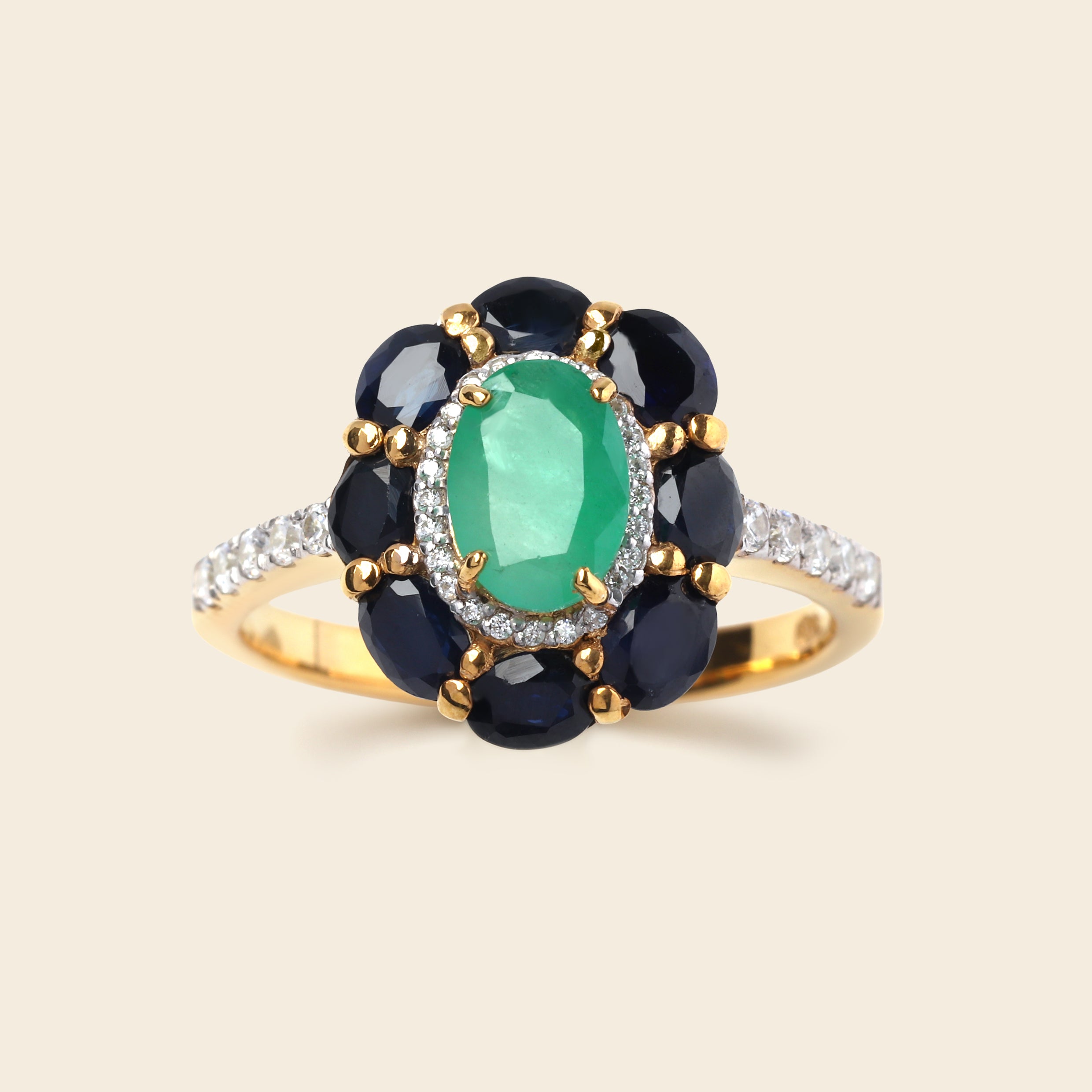 A yellow gold ring featuring a central oval green emerald-cut gemstone surrounded by a halo of dark blue round gemstones, with a pavé band of small clear stones on a light beige background.