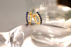 A pair of gold huggie hoop earrings embellished with small round blue gemstones, displayed on a reflective clear glass surface.
