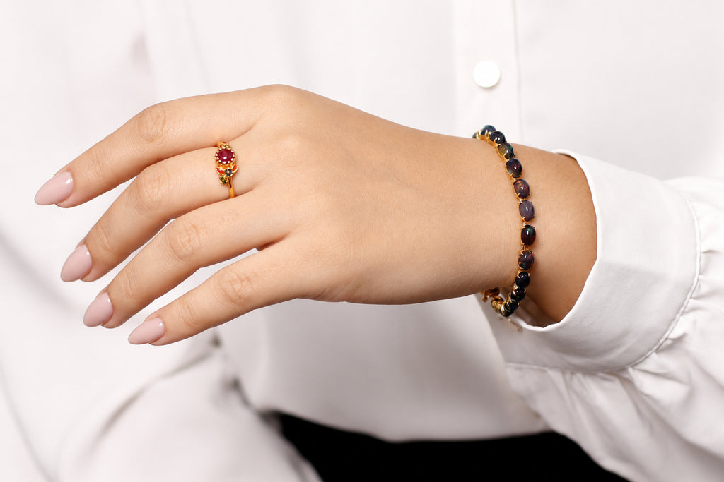A close-up of a person's hand wearing a gold ring with a red gemstone and a matching beaded bracelet on the wrist, styled with a white collared shirt.