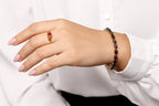 A close-up of a person's hand wearing a gold ring with a red gemstone and a matching beaded bracelet on the wrist, styled with a white collared shirt.