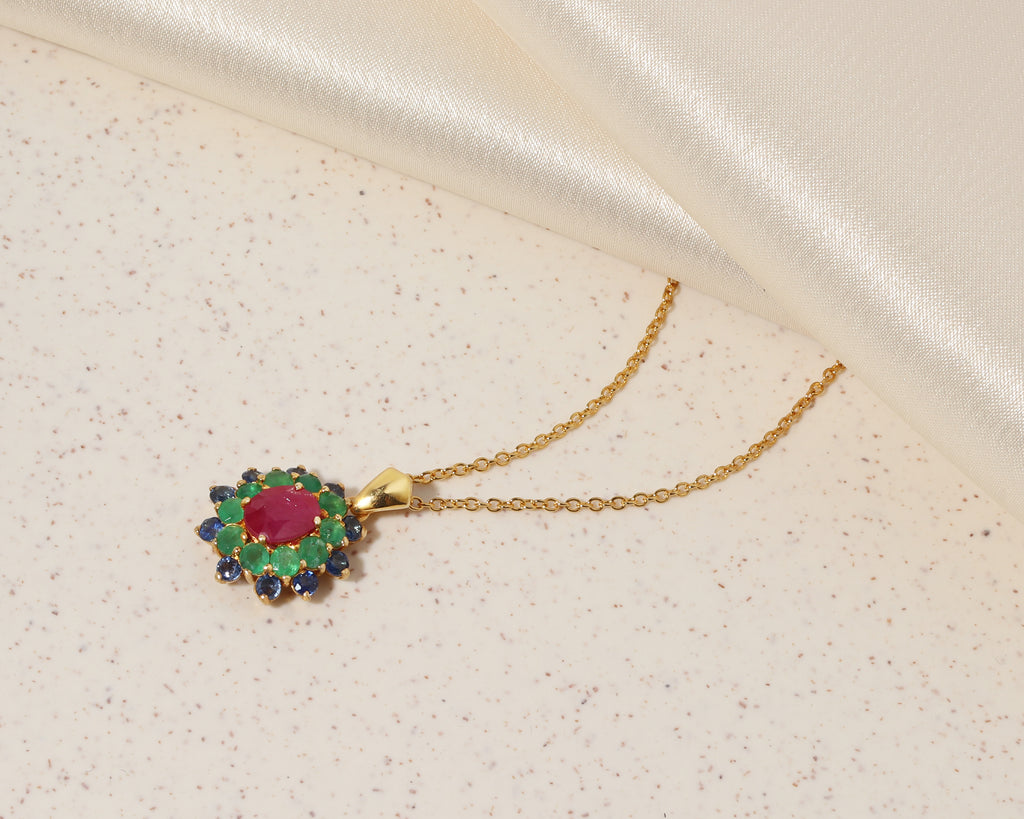A gold pendant necklace featuring a central oval red gemstone surrounded by green and dark blue stones, displayed on a textured cream surface with a satin fabric drape in the background.