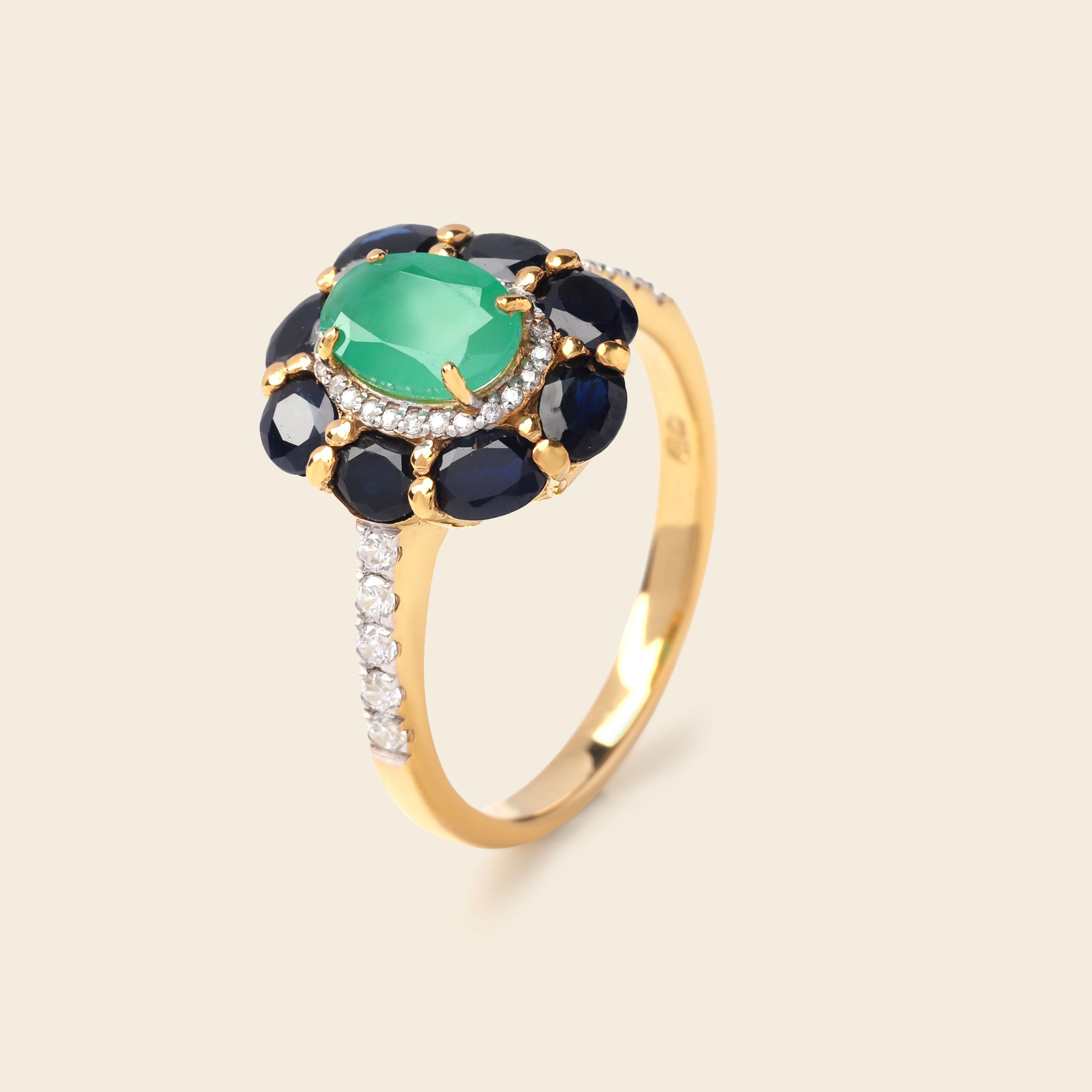 A yellow gold ring featuring a central green cushion-cut gemstone surrounded by a halo of dark blue oval gemstones and small clear accent stones, with a slim pavé-set band, shown on a light beige background.