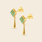 A pair of gold dangle earrings featuring a small cluster of green stones in a floral shape at the top, with a teardrop-shaped gold drop hanging below, set against a solid light beige background.