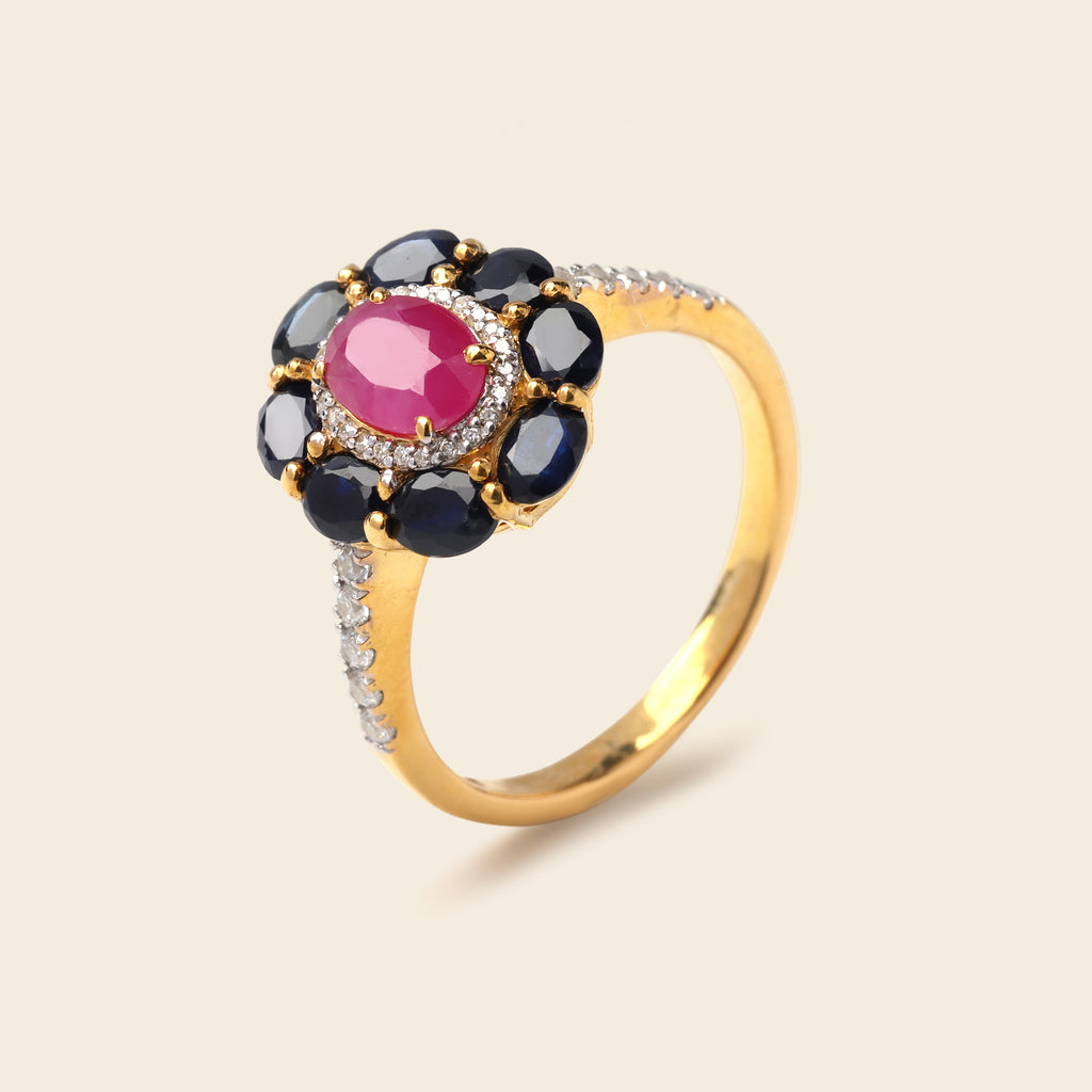 A gold ring featuring a central oval pink gemstone surrounded by a halo of round dark blue gemstones, accented with small clear stones on the band, set against a light beige background.