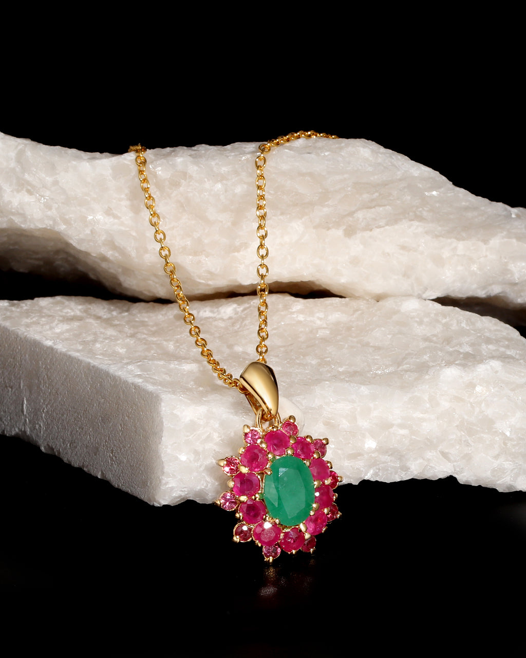 A gold pendant necklace featuring a central oval green gemstone surrounded by a halo of round pink gemstones, displayed on a white textured stone prop against a black background.