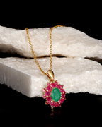 A gold pendant necklace featuring a central oval green gemstone surrounded by a halo of round pink gemstones, displayed on a white textured stone prop against a black background.