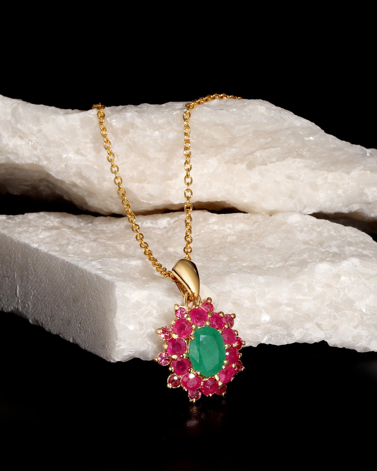 A gold pendant necklace featuring a central oval green gemstone surrounded by a halo of round pink gemstones, displayed on a white textured stone prop against a black background.