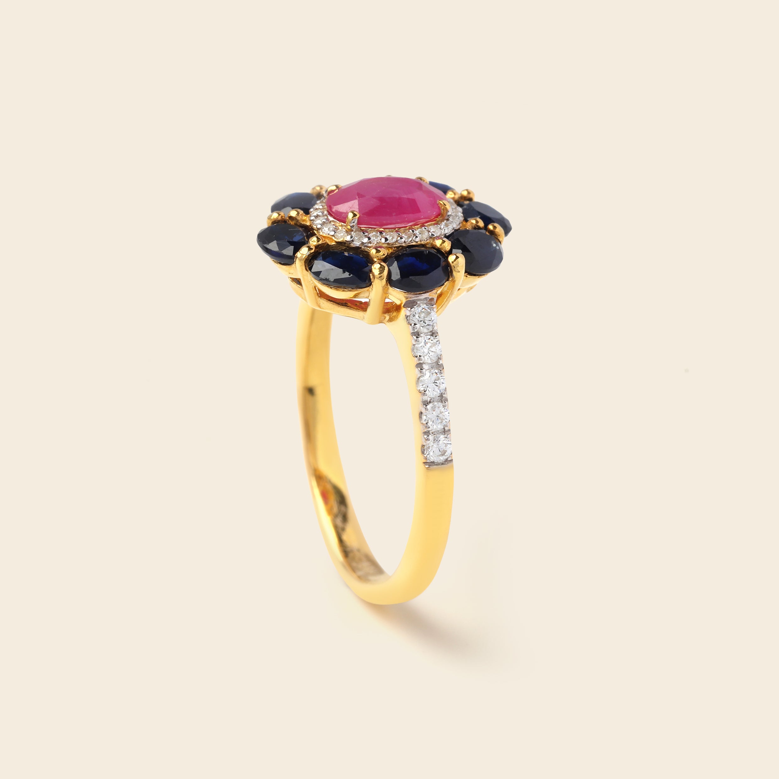 A yellow gold ring featuring a central oval pink gemstone surrounded by a halo of dark blue oval gemstones, with a pavé band set with small clear stones, shown on a light beige background.