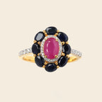 A yellow gold ring featuring a central oval pink gemstone surrounded by a halo of dark blue oval gemstones, with small clear accent stones on the band, set against a solid light beige background.