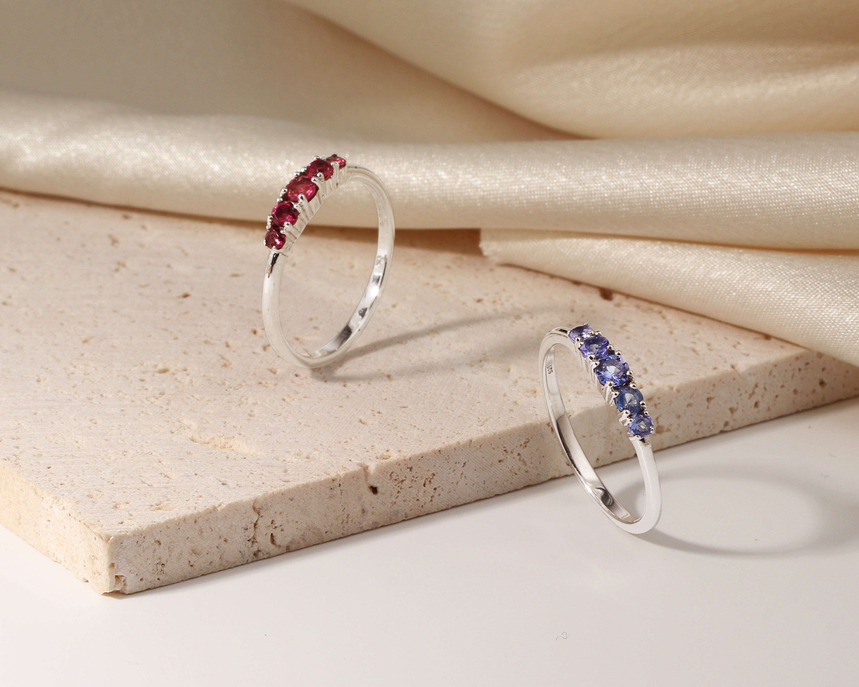 Two slim silver rings displayed on a beige stone slab with draped cream satin fabric in the background.