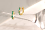 A pair of gold huggie hoop earrings adorned with a row of round-cut green gemstones, displayed on a reflective, light-colored surface.