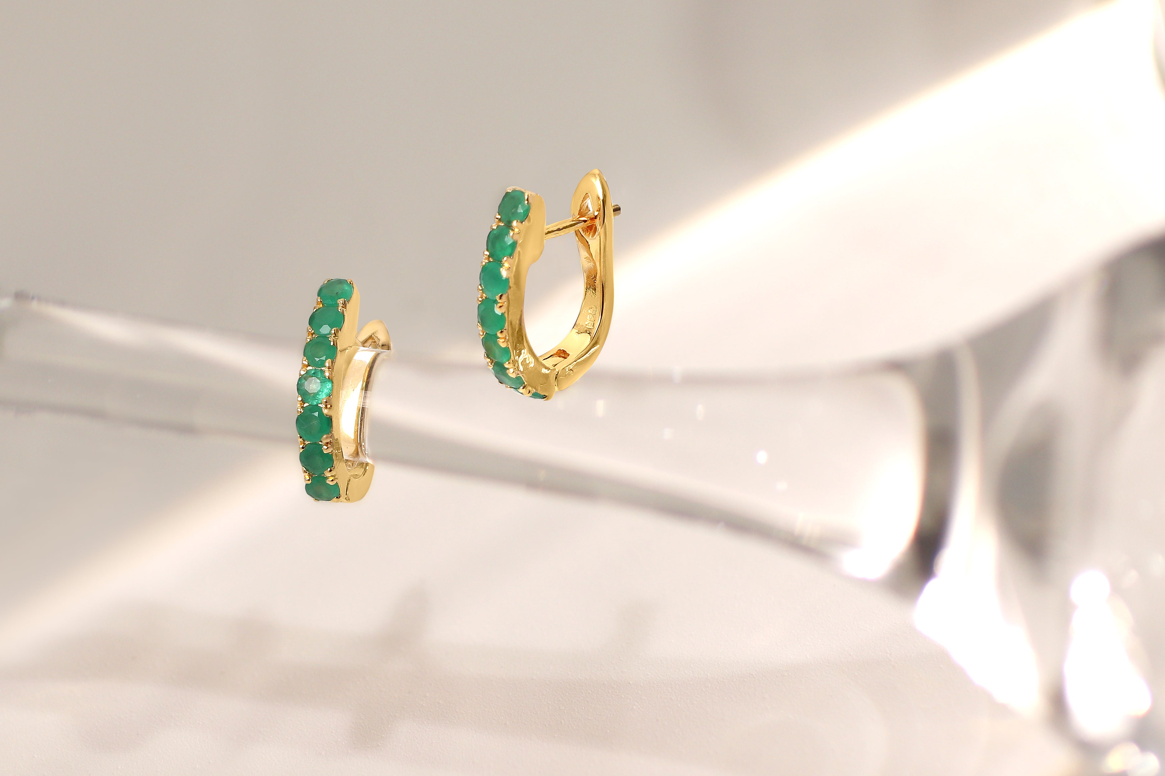 A pair of gold huggie hoop earrings adorned with a row of round-cut green gemstones, displayed on a reflective, light-colored surface.