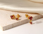 A pair of gold stud earrings with oval red gemstones, displayed on a textured beige stone slab with draped cream satin fabric in the background.