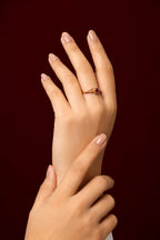 A close-up of a hand wearing a delicate gold ring with a small red gemstone, posed against a solid dark burgundy background.