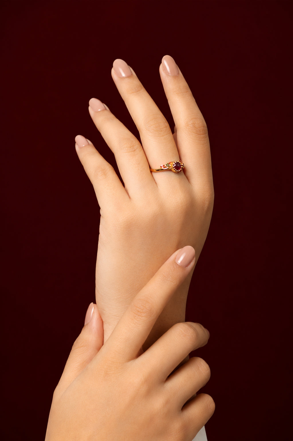 A close-up of a hand wearing a delicate gold ring with a small red gemstone, posed against a solid dark burgundy background.