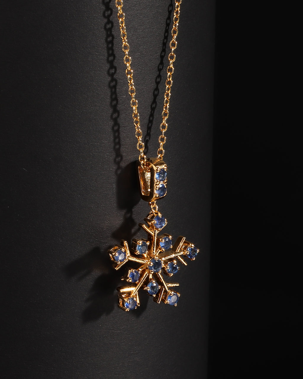 A gold snowflake pendant necklace adorned with small blue gemstones, displayed against a dark black background.