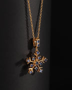 A gold snowflake pendant necklace adorned with small blue gemstones, displayed against a dark black background.