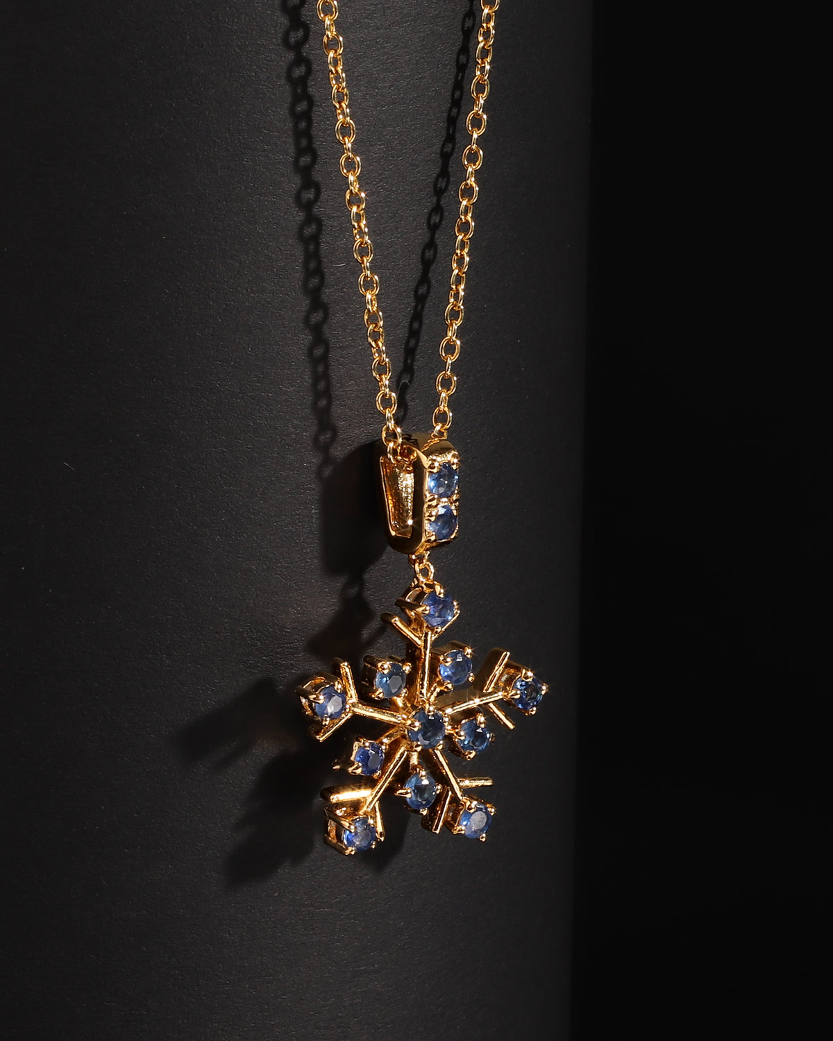A gold snowflake pendant necklace adorned with small blue gemstones, displayed against a dark black background.