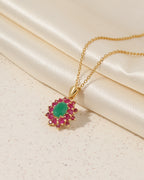 A gold pendant necklace featuring a central green oval stone surrounded by small pink gemstones in a gold setting, displayed on a cream satin fabric background.