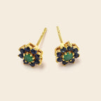 A pair of gold stud earrings featuring a central round green stone surrounded by a halo of small dark blue stones, set against a solid light beige background.