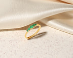 A gold ring featuring five small round green gemstones set across the top, displayed on a textured cream surface with draped satin fabric in the background.