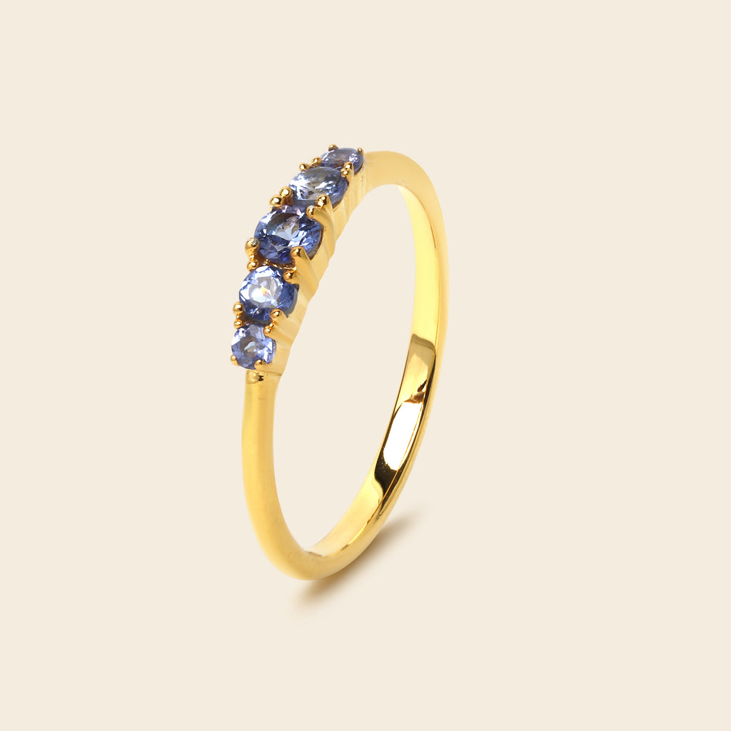 A yellow gold ring featuring a row of five round-cut blue gemstones, set against a solid light beige background.