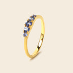A yellow gold ring featuring a row of five round-cut blue gemstones, set against a solid light beige background.
