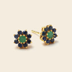 A pair of gold flower-shaped stud earrings featuring a central green stone surrounded by a halo of dark blue stones, set against a light beige background.
