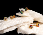 Four gold flower-shaped stud earrings with multicolored gemstones are displayed on white textured stone blocks against a solid black background.