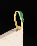 A yellow gold ring with a row of green emerald-cut gemstones, displayed on a textured white block against a solid black background.