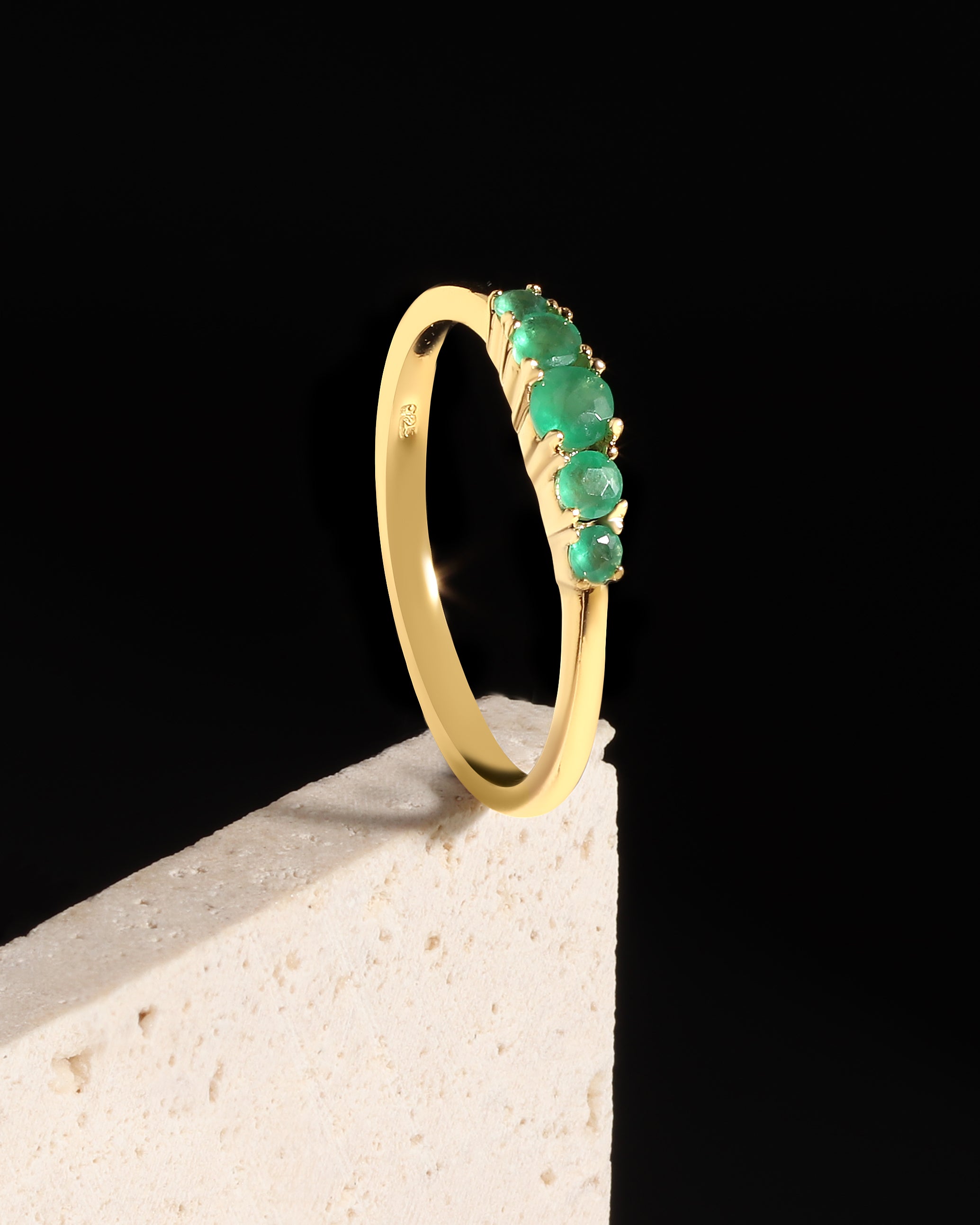 A yellow gold ring with a row of green emerald-cut gemstones, displayed on a textured white block against a solid black background.