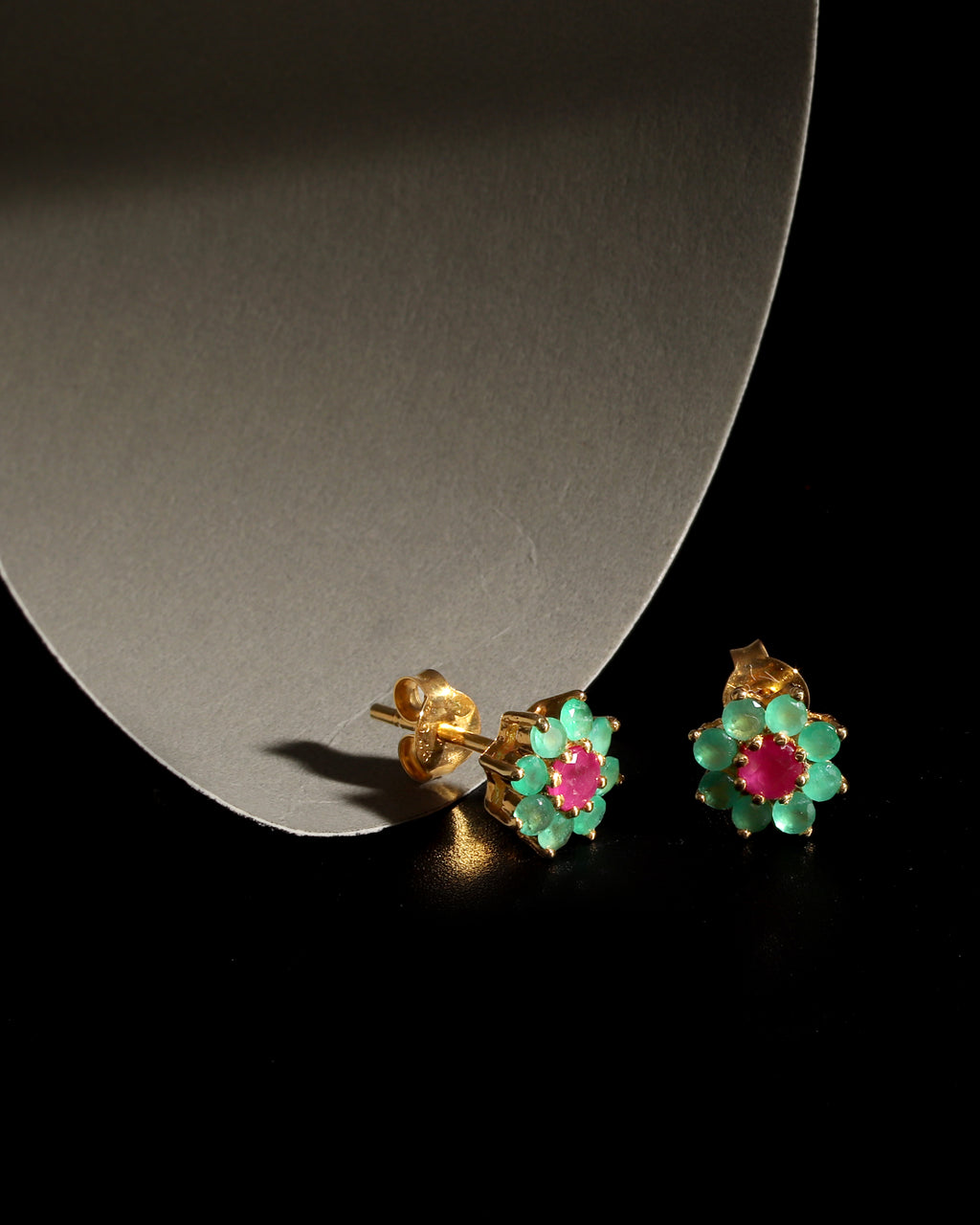 A pair of gold flower-shaped stud earrings with green enamel petals and pink centers, displayed on a black surface next to a curved gray stand.