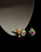A pair of gold flower-shaped stud earrings with green enamel petals and pink centers, displayed on a black surface next to a curved gray stand.